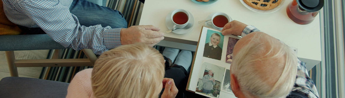 Drie oudere personen zitten rond een tafel met koffie, koekjes en taart. Op tafel ligt een fotoalbum dat samen wordt bekeken. De foto's tonen herinneringen uit verschillende levensfasen. De setting straalt rust uit en illustreert hoe herinneringen en verhalen een plaats kunnen krijgen in het dagelijks leven.