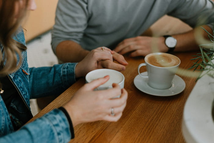 Twee personen in gesprek aan een houten tafel met koffie in een gezellige caféomgeving.