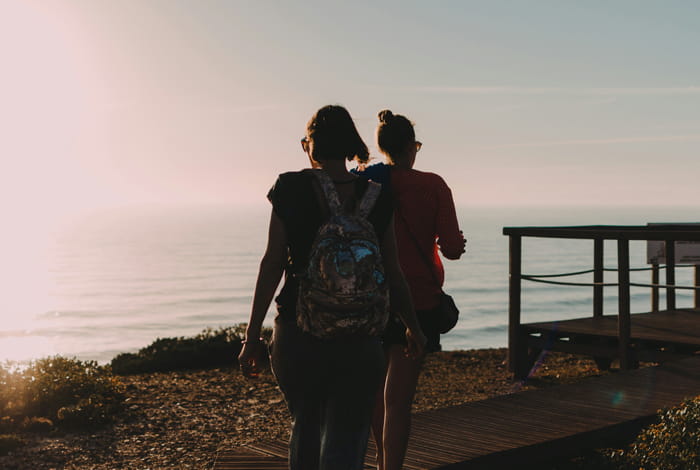 Twee personen wandelen langs een houten pad met uitzicht op zee tijdens zonsondergang.