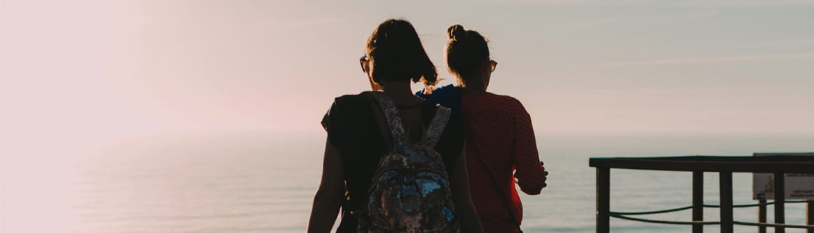 Twee personen wandelen langs een houten pad met uitzicht op zee tijdens zonsondergang.
