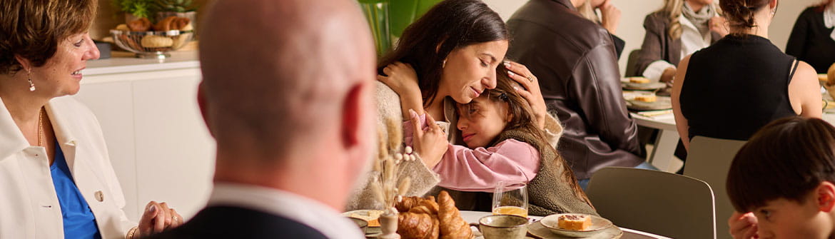 Un groupe de personnes est assis autour d’une table dressée avec des pains, des douceurs, des boissons et de la vaisselle lors d’un repas partagé.