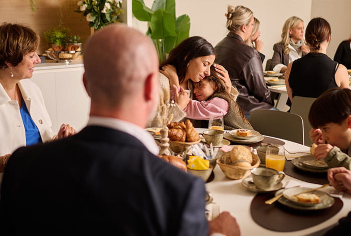 Un groupe de personnes est assis autour d’une table dressée avec des pains, des douceurs, des boissons et de la vaisselle lors d’un repas partagé.