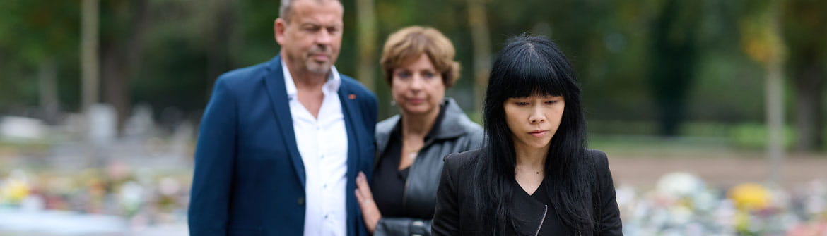 Drie personen staan bij een kist met bloemen op een begraafplaats, omringd door groen en grafstenen.
