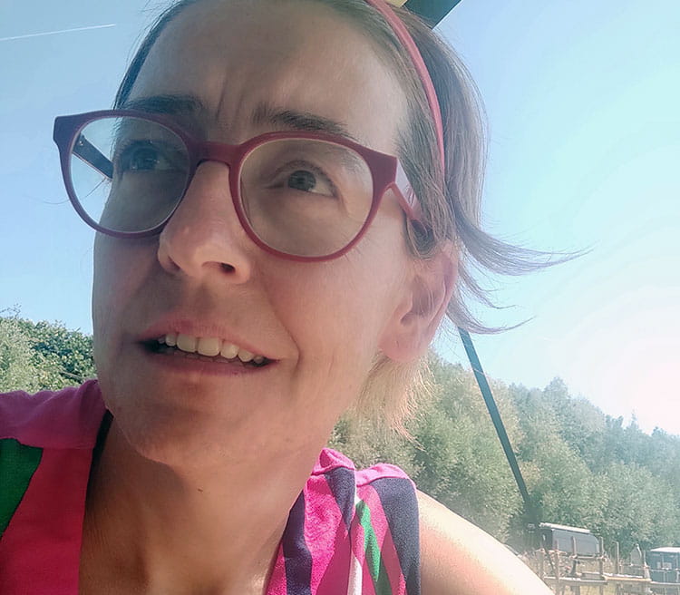 Close-up van een persoon met ronde bril en een haarband, buiten gefotografeerd onder een zonnescherm, met een landelijke omgeving op de achtergrond.