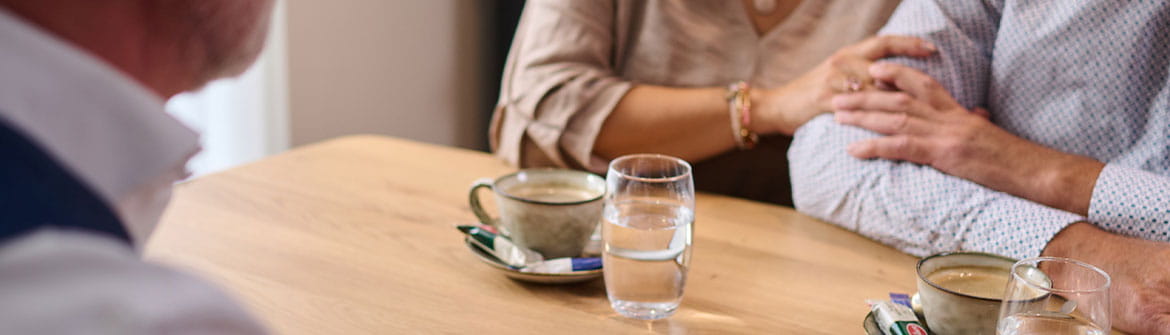 Deux personnes sont assises à une table en entretien avec un collaborateur funéraire. Sur la table se trouvent des documents, des tasses de café et des verres d’eau, tandis qu’une main repose en signe de soutien sur un bras. 