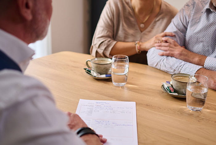 Twee personen zitten aan een tafel in gesprek met een uitvaartmedewerker. Op tafel zie je documenten, koffiekopjes en glazen water, terwijl een hand steunend op een arm rust.
