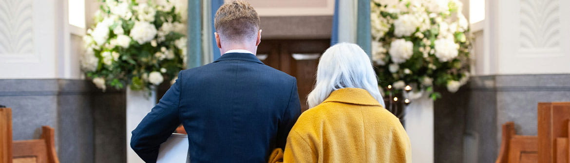 Deux personnes marchent ensemble dans une salle de cérémonie funéraire, entourée de bancs en bois et de grands arrangements floraux.
