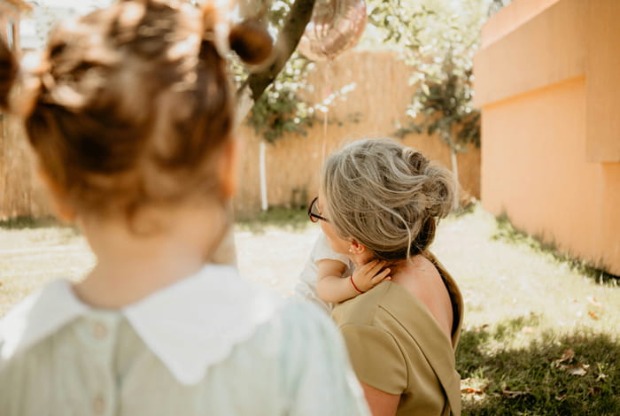 Un adulte tient un bébé dans ses bras dans un jardin ensoleillé, tandis qu'un jeune enfant se tient au premier plan.