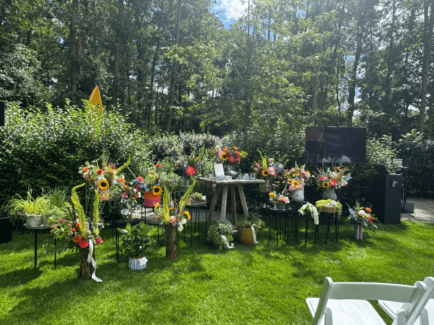 Lieu de cérémonie d’adieu aménagé dans un jardin verdoyant. De nombreuses compositions florales colorées sont disposées autour d’une table commémorative centrale. À droite, un écran et une installation sonore sont prévus pour une cérémonie funéraire en plein air. Des chaises blanches pour les participants sont visibles au premier plan. En arrière-plan, des arbres et des arbustes créent un cadre naturel et apaisant.