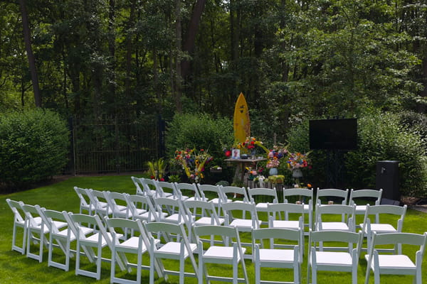 Des rangées de chaises blanches sont installées sur une pelouse dans un cadre boisé pour une cérémonie d’adieu en plein air. À l’avant se trouve un espace commémoratif avec des compositions florales colorées, des objets personnels et une photo.