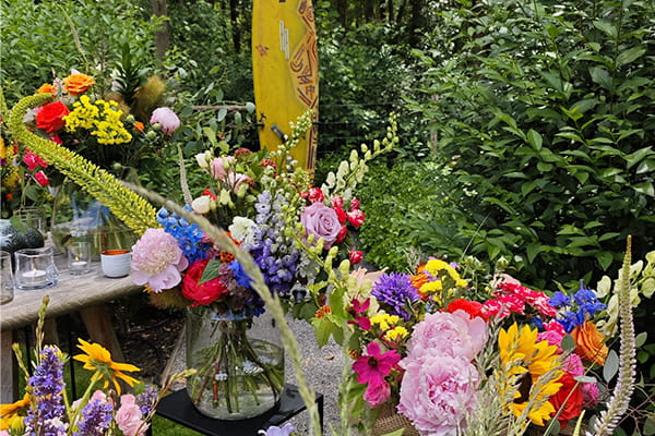 Des compositions florales colorées et des fleurs des champs créent un espace commémoratif personnalisé dans un cadre verdoyant. Parmi les fleurs se dresse une planche de surf jaune, utilisée comme hommage marquant lors d’une cérémonie d’adieu en plein air.