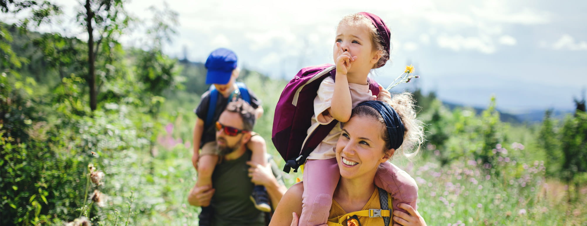 Twee volwassenen wandelen met twee kinderen op een natuurpad; één kind zit op de schouders en draagt een rugzak, terwijl het andere kind ook gedragen wordt.