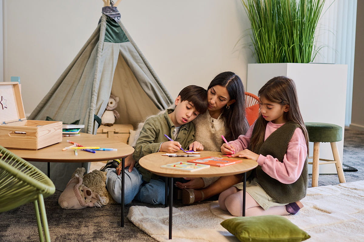 Volwassene en twee kinderen kleuren samen aan lage tafeltjes in een kindvriendelijke ruimte.