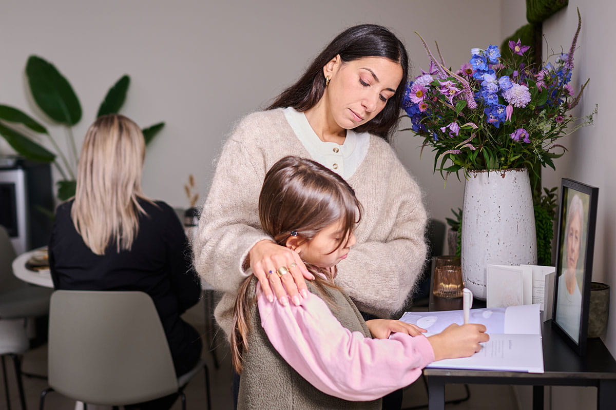 Un adulte accompagne un enfant qui écrit un message dans un livre de condoléances, à côté de fleurs et d’un portrait.