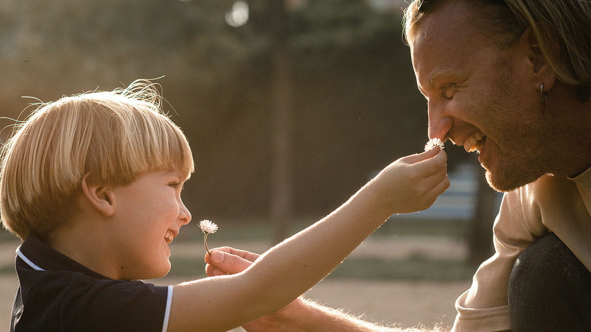 Un enfant et un adulte sont assis l'un en face de l'autre à l'extérieur. L'enfant tient une fleur et en tend une autre à l'adulte.