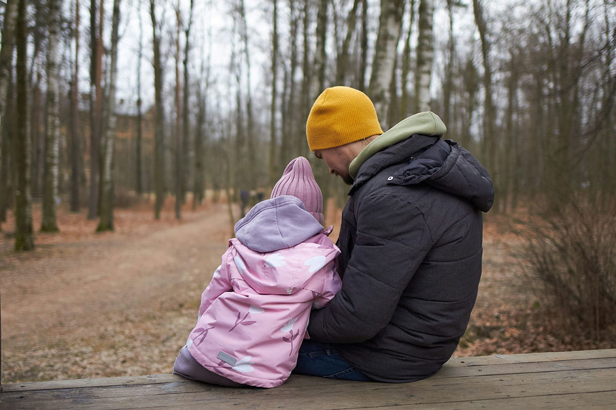 Un adulte et un enfant sont assis côte à côte sur un banc en bois dans un environnement naturel boisé. Ils regardent un sentier entre les arbres tandis que l’adulte entoure l’enfant de son bras.