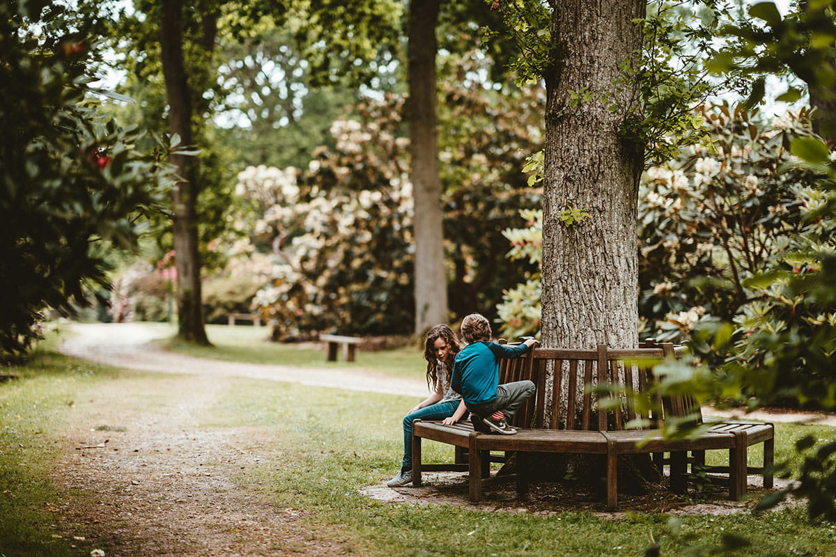 Twee kinderen zitten op een houten zitbank rond een boom in een park met bomen en wandelpaden op de achtergrond.