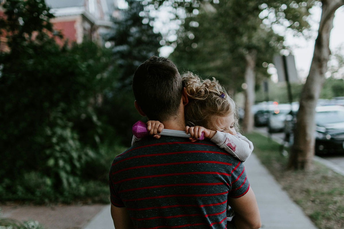 Un adulte porte un jeune enfant dans ses bras sur un chemin bordé d’arbres.
