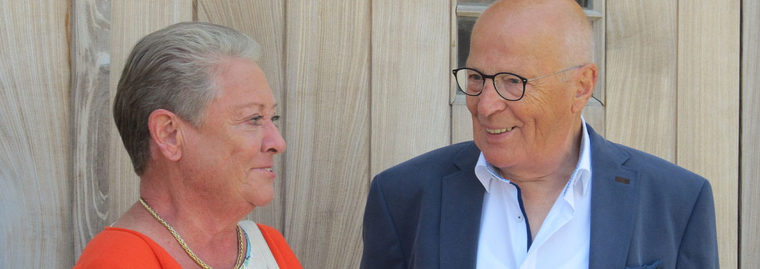 Une femme âgée et un homme âgé posent ensemble devant une porte en bois, vêtus de tenues élégantes lors d’une occasion spéciale.