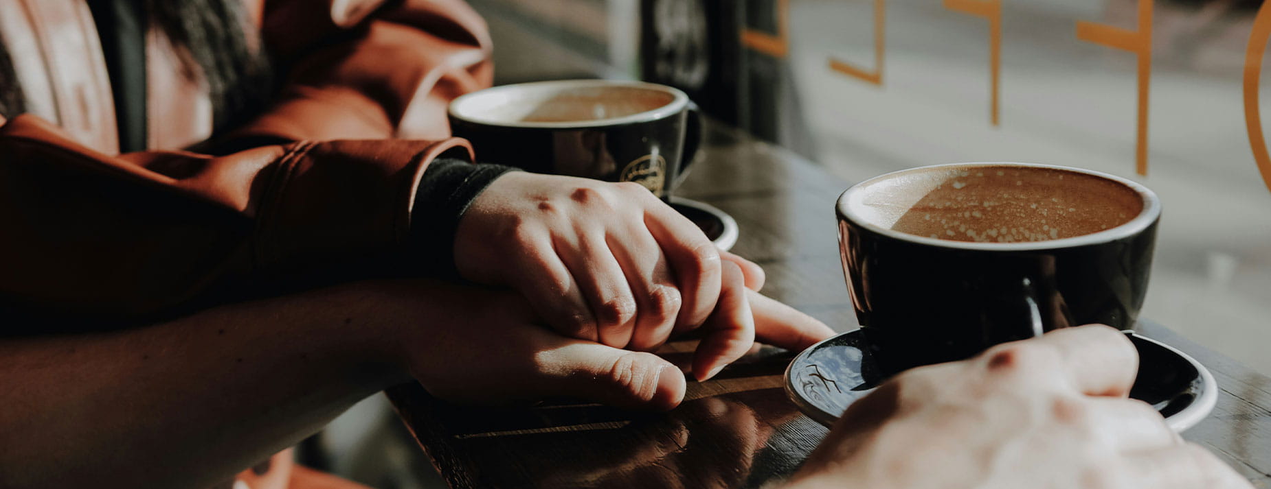 Deux personnes sont assises à une table près d’une fenêtre, chacune avec une tasse de café. Une main repose en signe de soutien sur celle de l’autre pendant une conversation.