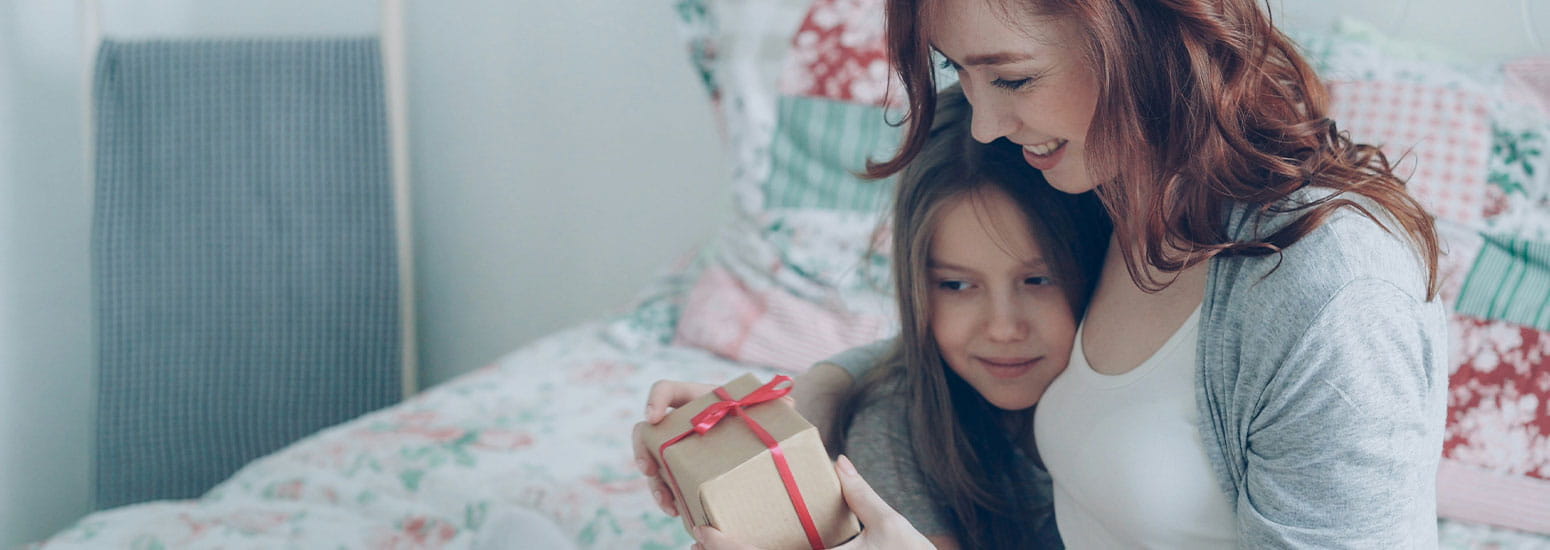 Deux personnes sont assises ensemble sur un lit tandis que l’une d’elles tient un petit cadeau emballé avec un ruban rouge. La chambre présente des tons doux et un lit avec une housse à motif floral en arrière-plan.