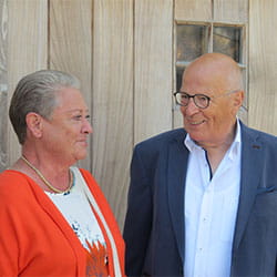 Une femme âgée et un homme âgé posent ensemble devant une porte en bois, vêtus de tenues élégantes lors d’une occasion spéciale.