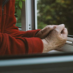 Gros plan de deux mains jointes posées sur un rebord de fenêtre, avec un vêtement rouge et une nature verdoyante en arrière‑plan.