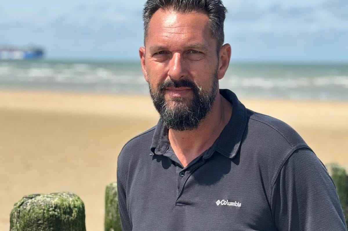 Personne aux cheveux courts foncés et barbe, debout près de poteaux en bois sur la plage, avec la mer et un bateau flou en arrière-plan.