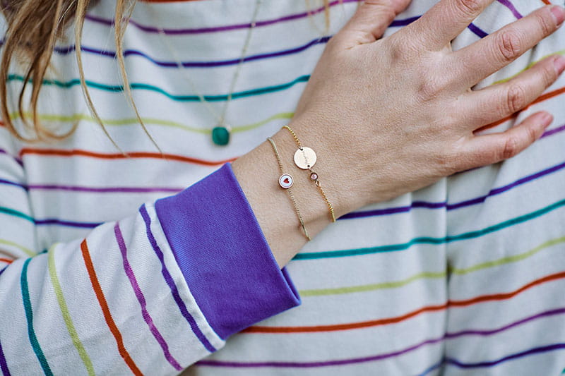 Close-up van een hand op de borst van een persoon die een gestreepte trui draagt, met twee fijne armbandjes om de pols, waarvan één met een klein hartje.