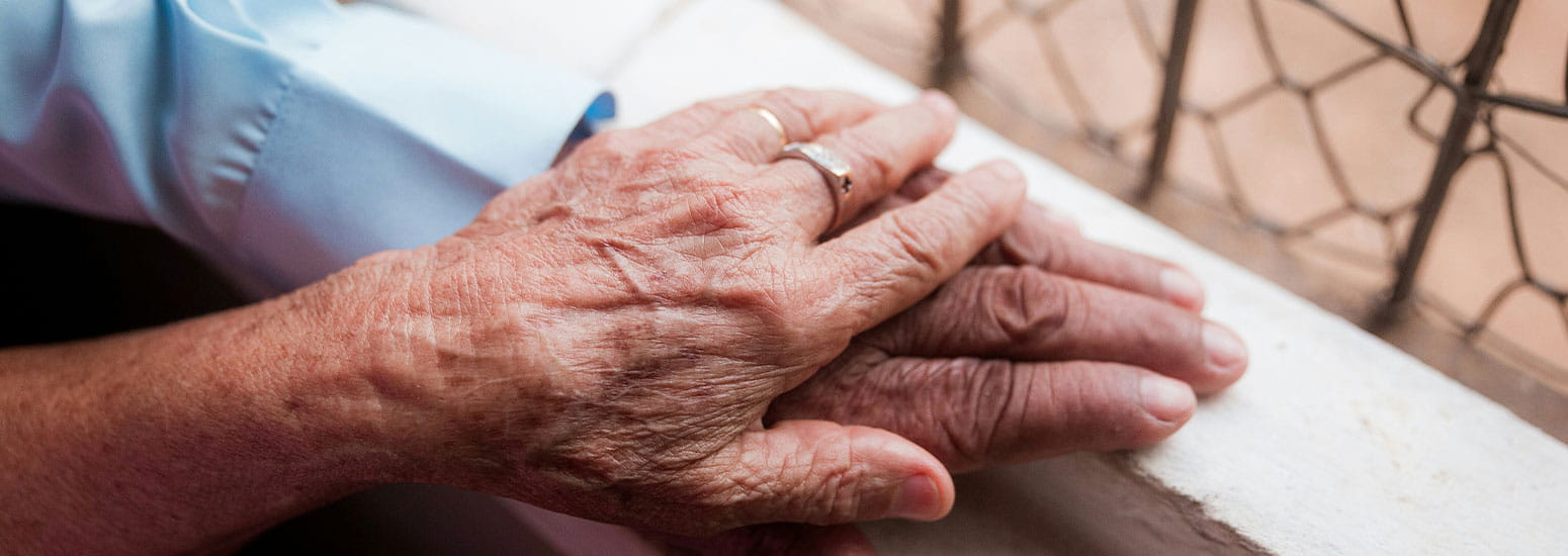 Close‑up van twee handen die op elkaar rusten op een vensterbank, met een hekwerk buiten het raam op de achtergrond. 