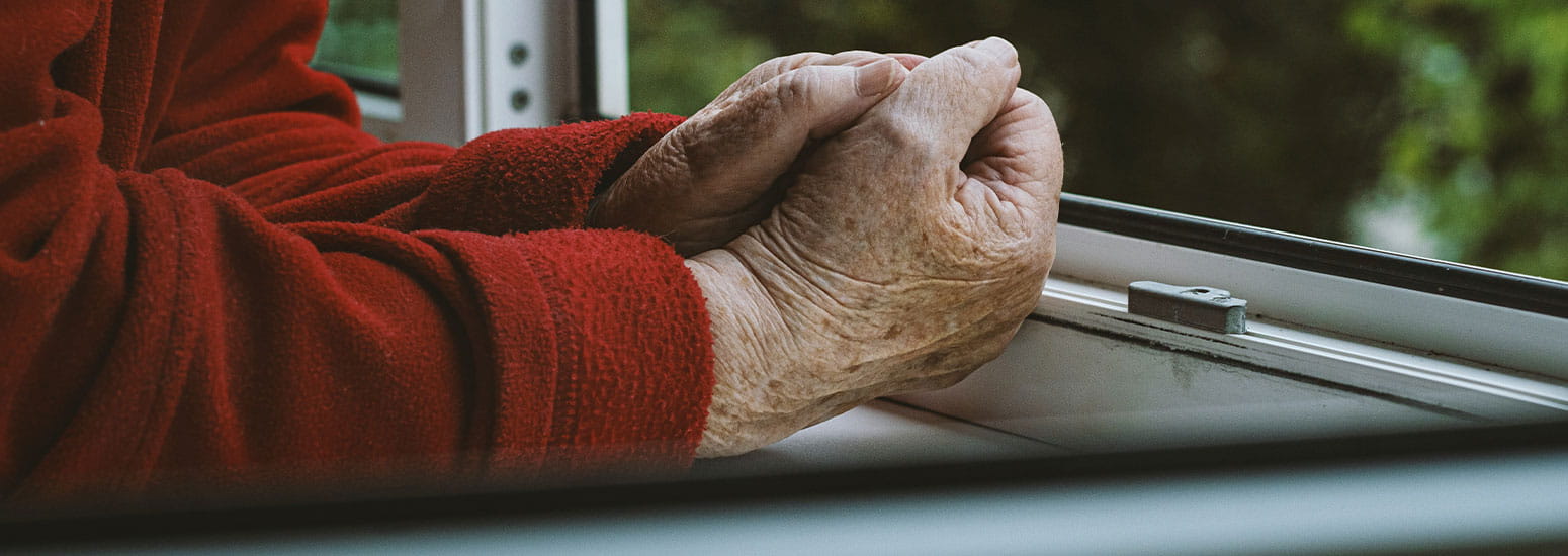 Close‑up van twee gevouwen handen op een vensterbank, met een rood kledingstuk zichtbaar en groene natuur op de achtergrond.